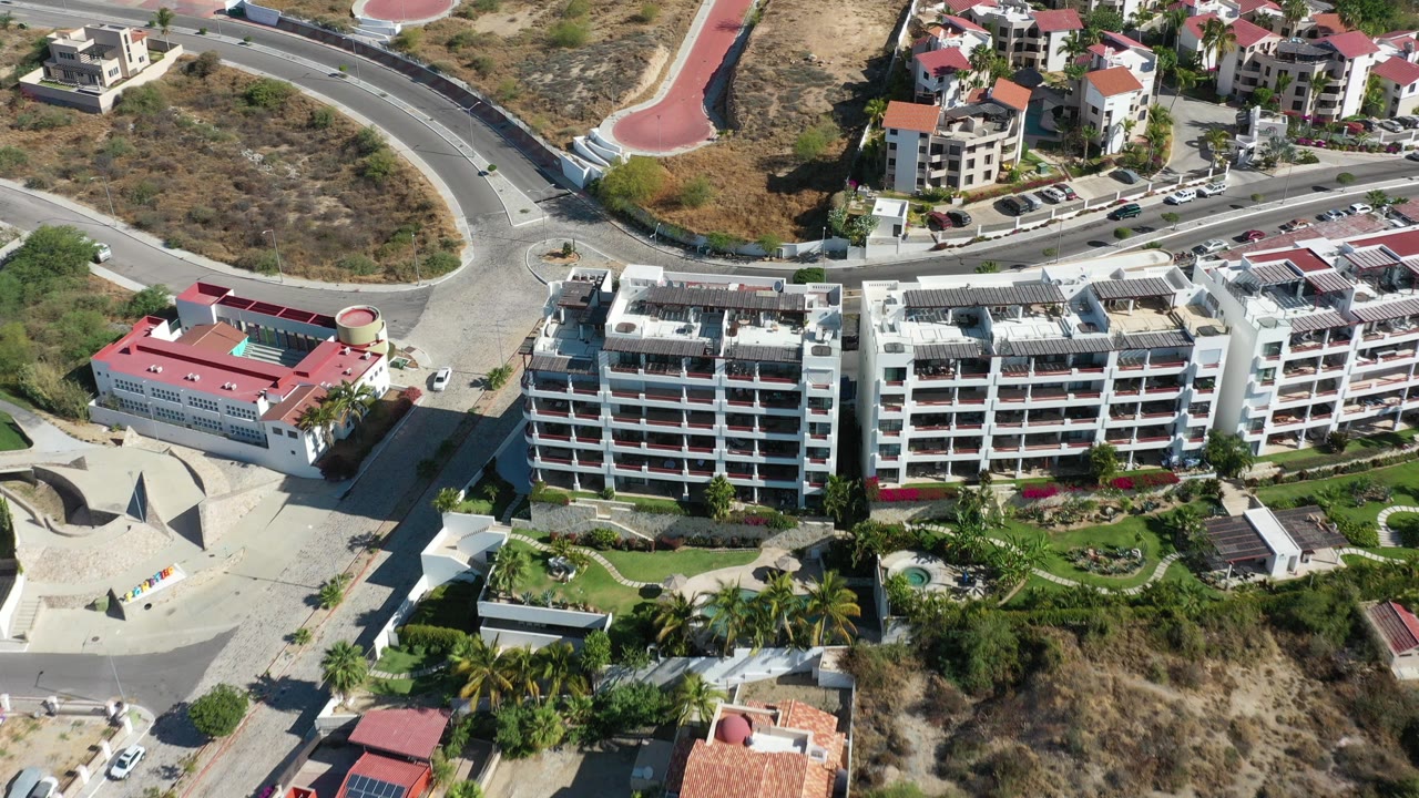 Oblique aerial view over Los Cabos, Mexico, with white multi-story buildings, red roofs, curved roads, parked cars, dry hillside, and no visible people.