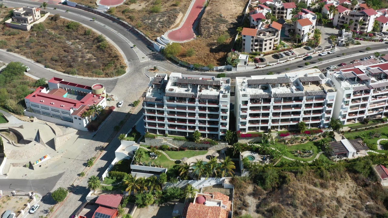 Aerial frame over Los Cabos, Mexico, showing apartment-style buildings, landscaped palms, a looping road, parked cars, and no visible people.