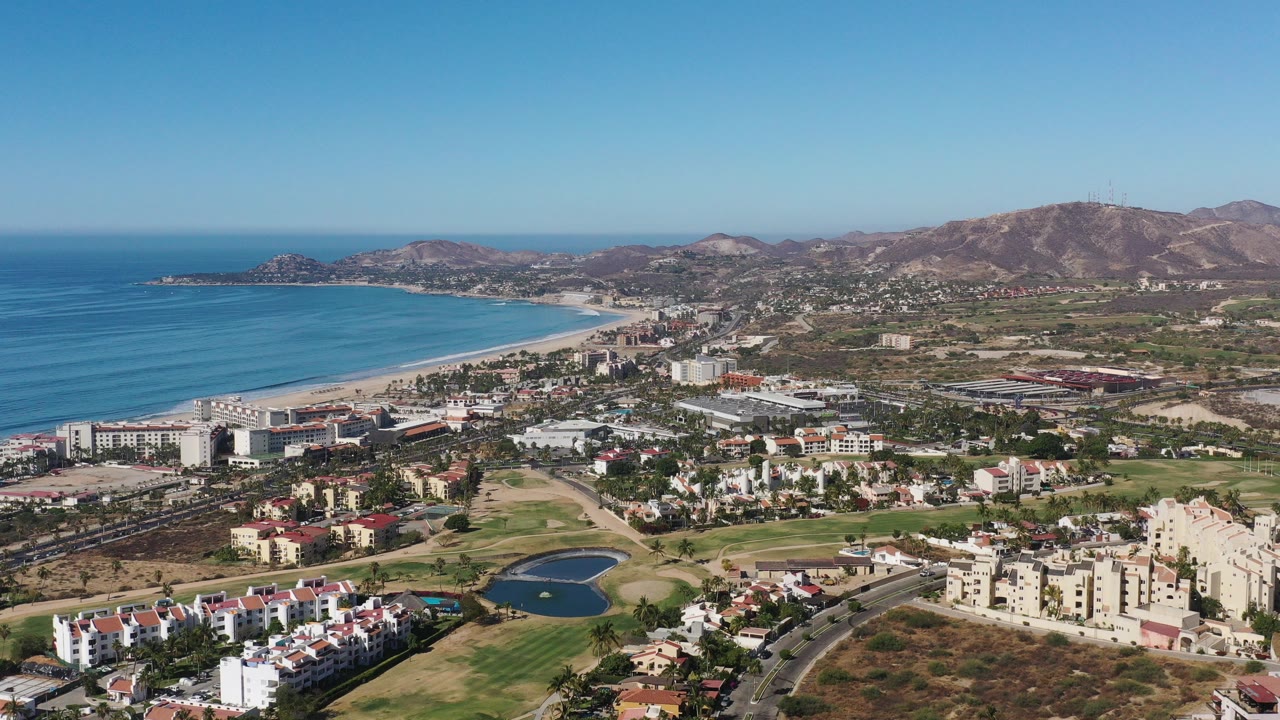 Wide aerial view in Los Cabos, Mexico, with blue ocean and beach at left, residential buildings and golf greens below, dry hills in the distance, and no visible people.