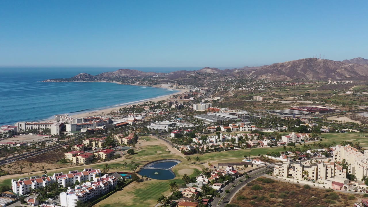High aerial view of ocean, beachside buildings, houses, green fairways, and mountains.