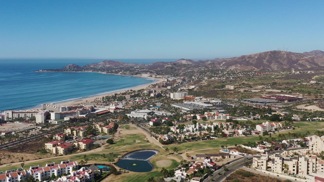Aerial view of coastline, town buildings, golf-course greenery, ponds, and dry hills.