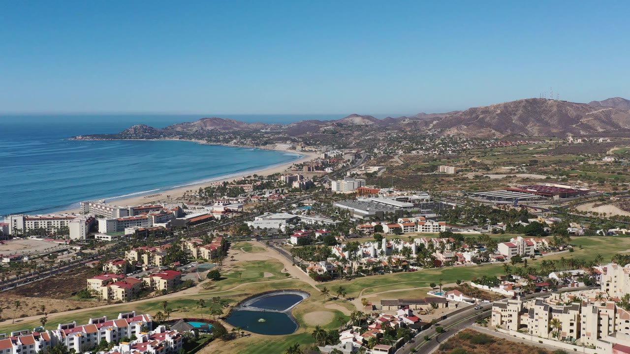 Aerial frame from Los Cabos, Mexico, showing the coastline stretching left to center, clustered buildings, green course areas, small ponds, brown hills, and no visible people.