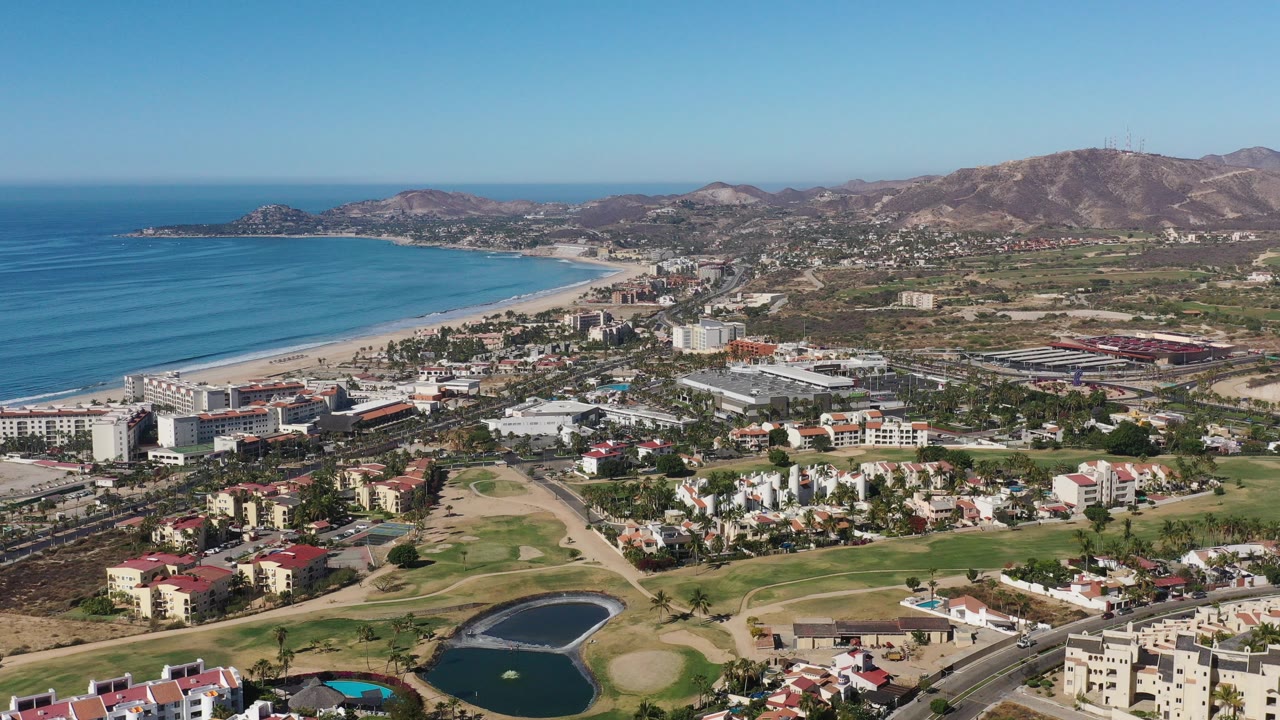 Aerial view of coastal buildings, roads, beach, ocean, and dry hills.
