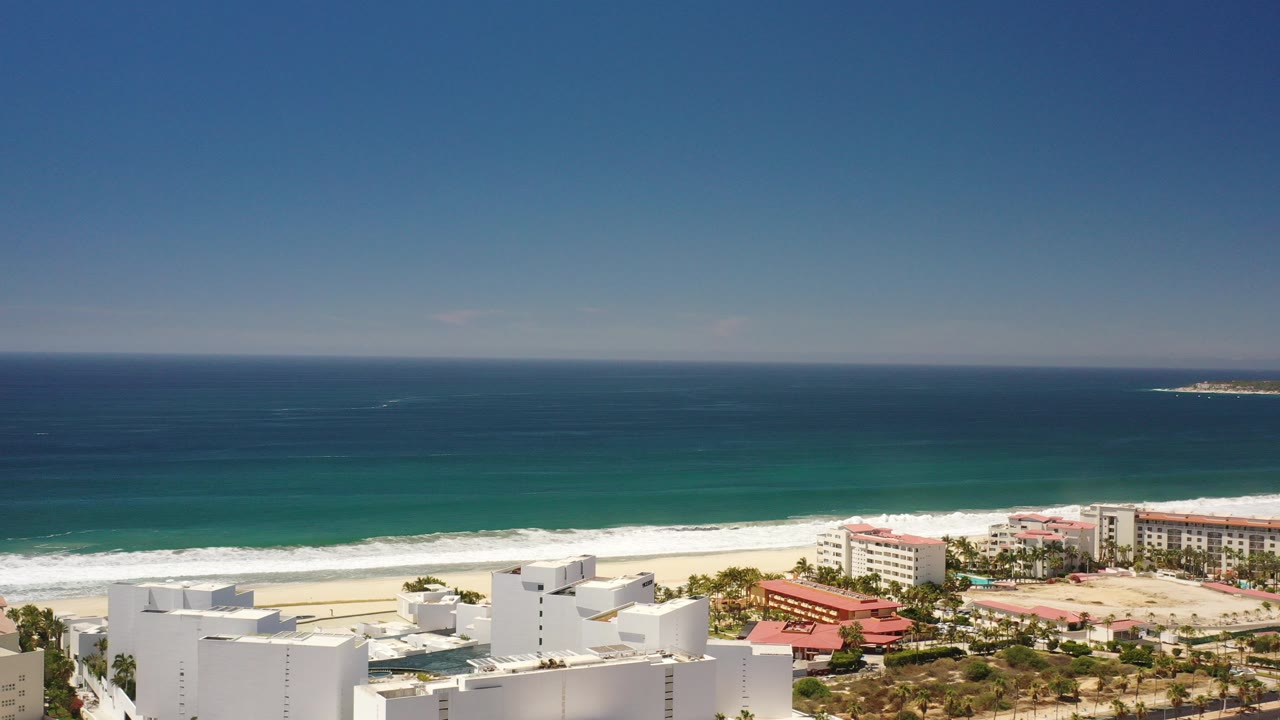 High aerial view of the Los Cabos coastline with deep blue ocean, turquoise shallows, pale sand, white beachfront buildings, red roofs, and no visible people.