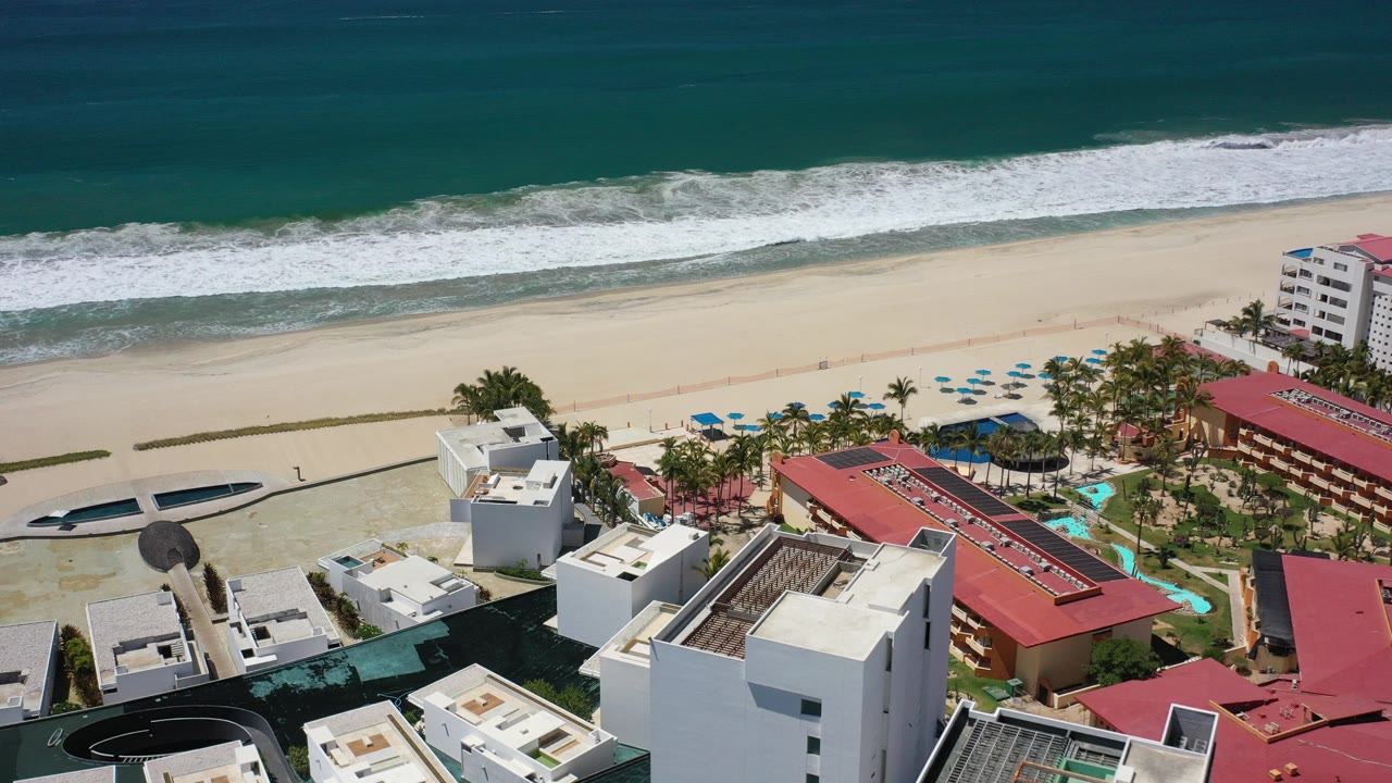 High aerial frame over Los Cabos beachfront buildings with white roofs, red roofs, palms, pale sand, blue-green ocean, and no visible people.