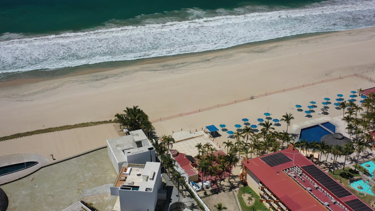 High aerial view in Los Cabos, Mexico with foamy surf at the top, wide sand below it, palm trees, blue umbrellas, pools, and beachfront buildings, with no visible people discernible.