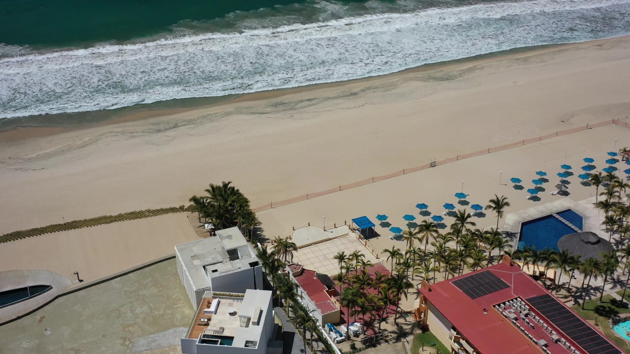 Oblique drone view of ocean waves, sandy beach, palm-lined property, and pool areas.