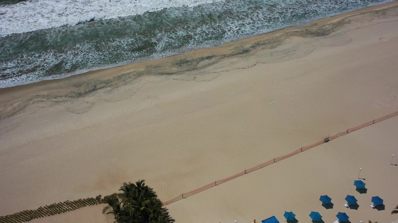 Aerial view over a Los Cabos beach with white surf at the top, broad tan sand, blue umbrellas near the lower edge, a red-orange barrier across the sand, and no visible people.