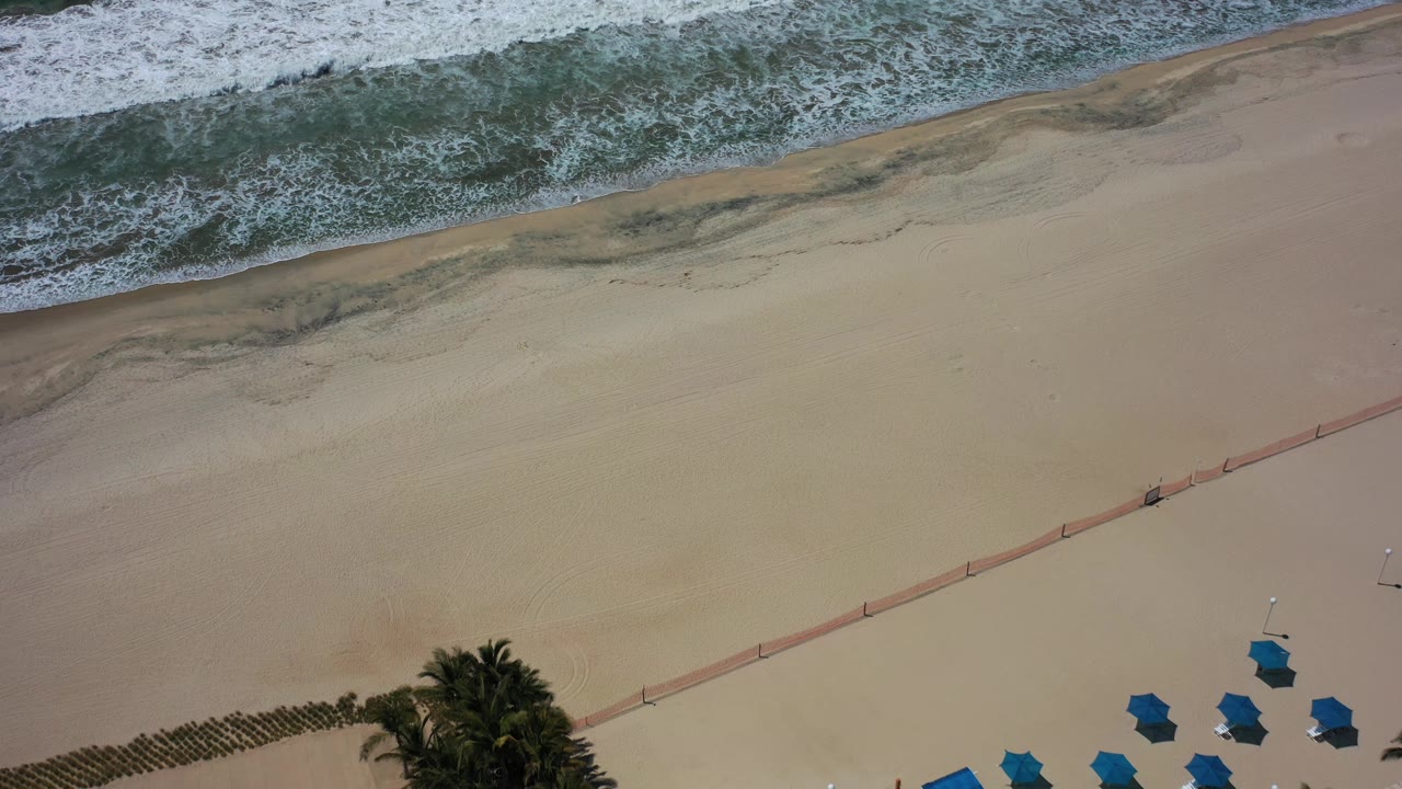 Aerial Los Cabos beach frame with foamy waves along the upper edge, open sand below, blue umbrellas grouped near the bottom, a diagonal red-orange barrier, and no visible people.