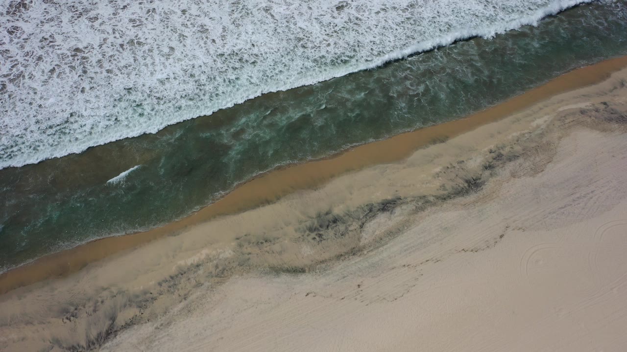 Top-down view of a Los Cabos shoreline where white foam, dark green water, wet black sand, and pale dry sand form diagonal bands, with no visible people.