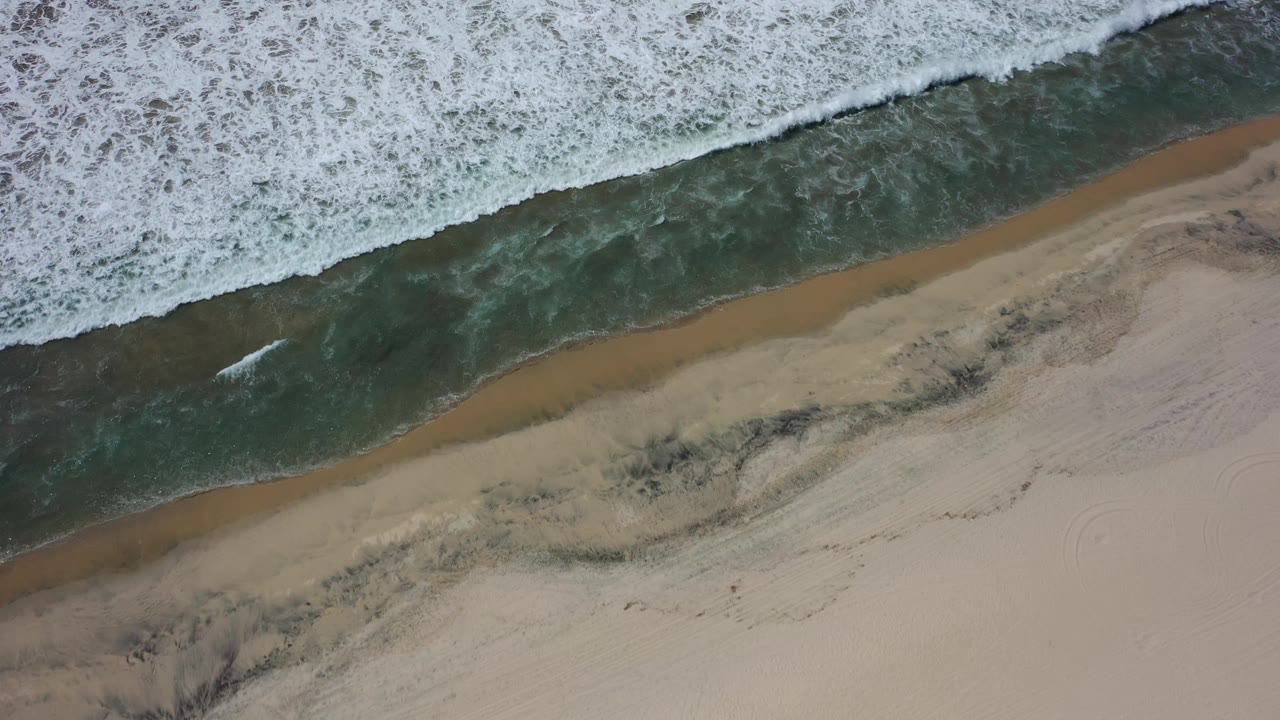 Aerial frame of Los Cabos surf with a broader white foam edge pressing toward the beach, leaving a narrow dark water band and tan sand below, with no visible people.