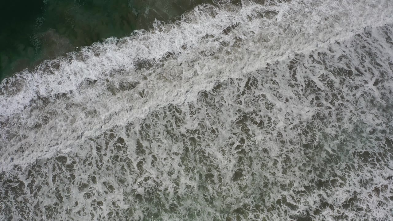 Overhead view in Los Cabos, Mexico, as the foamy wave band fills more of the frame over green water, with no visible people.