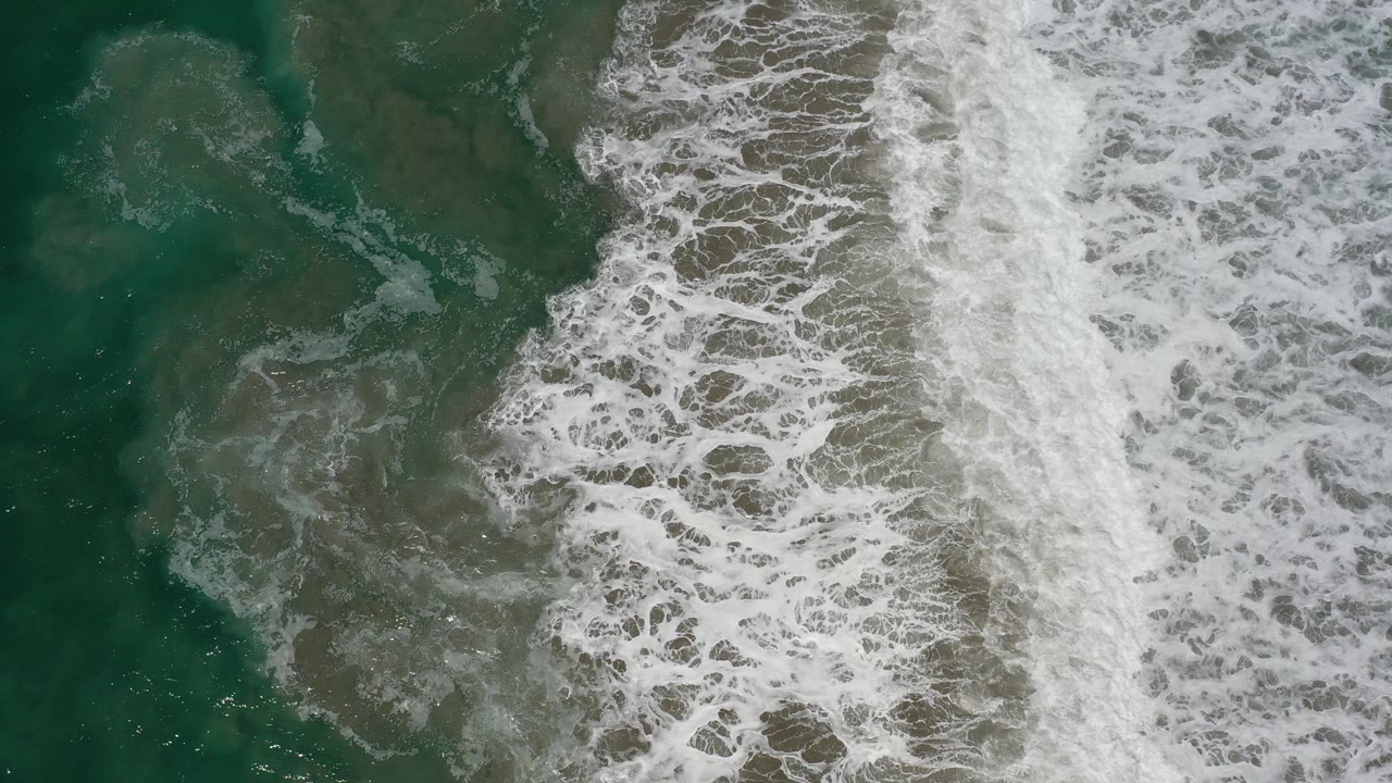 Overhead view of green ocean water in Los Cabos, Mexico, with a wide band of white surf foam entering from the right and no visible people.