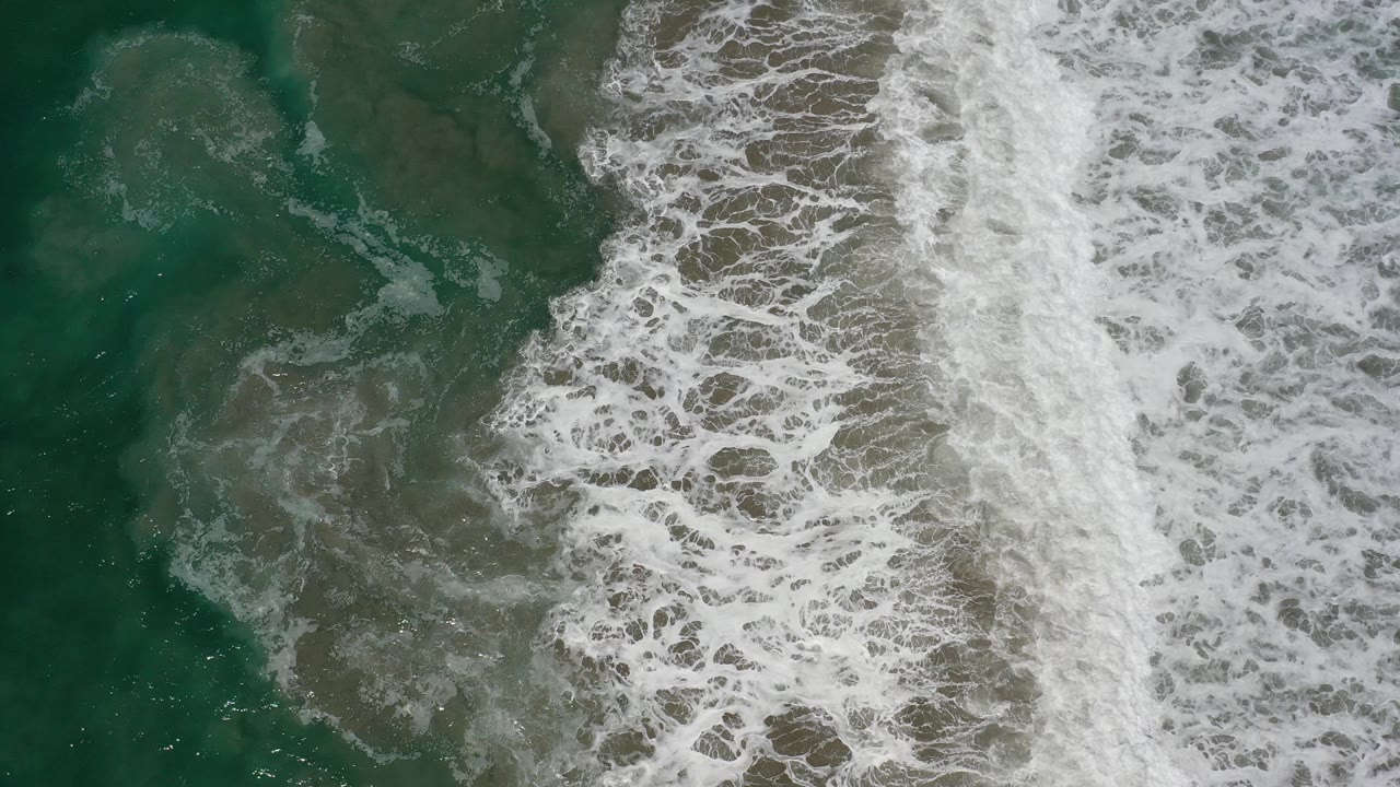 Aerial frame over Los Cabos ocean water where white foam spreads farther across the center, with darker green water still visible on the left and no visible people.