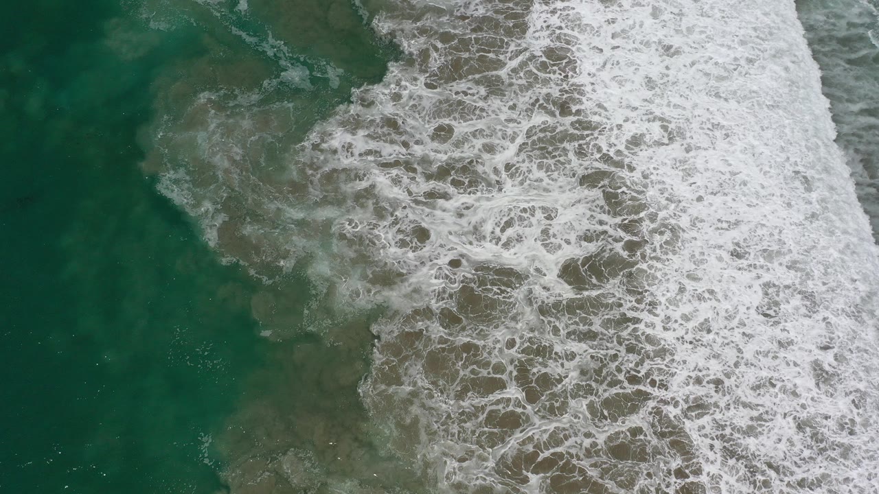 Overhead view in Los Cabos, Mexico, with green ocean water, white foam spreading over darker shallow areas, and no visible people.