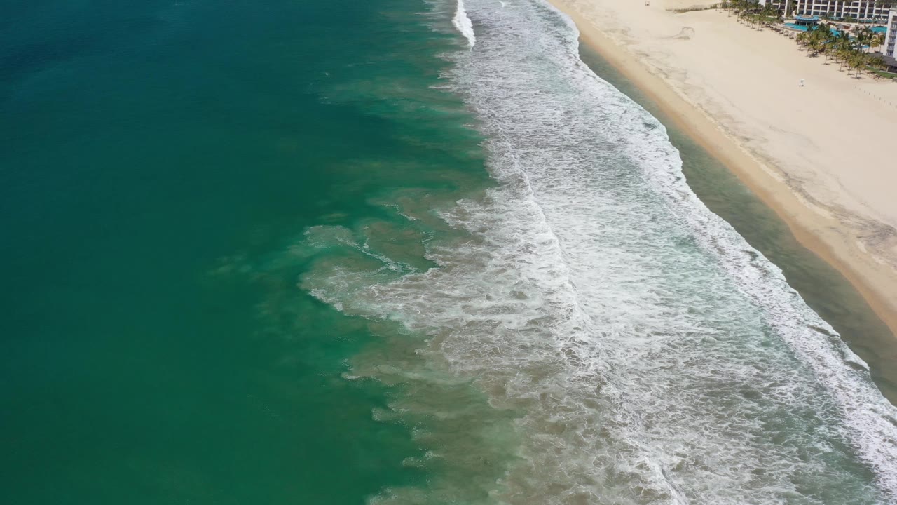Los Cabos aerial shoreline with a broad band of foamy white water near the beach and beachfront buildings beginning to appear at the upper right, with no visible people.