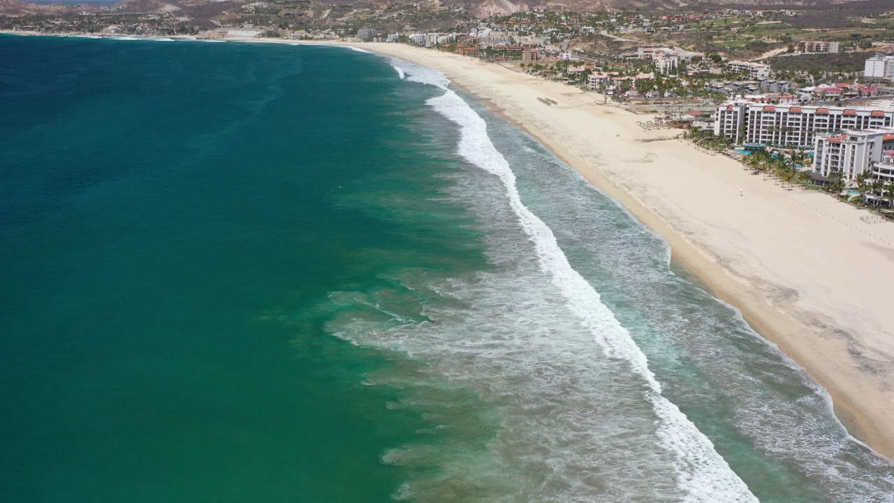 Aerial view of turquoise ocean waves reaching a wide beach with buildings inland.