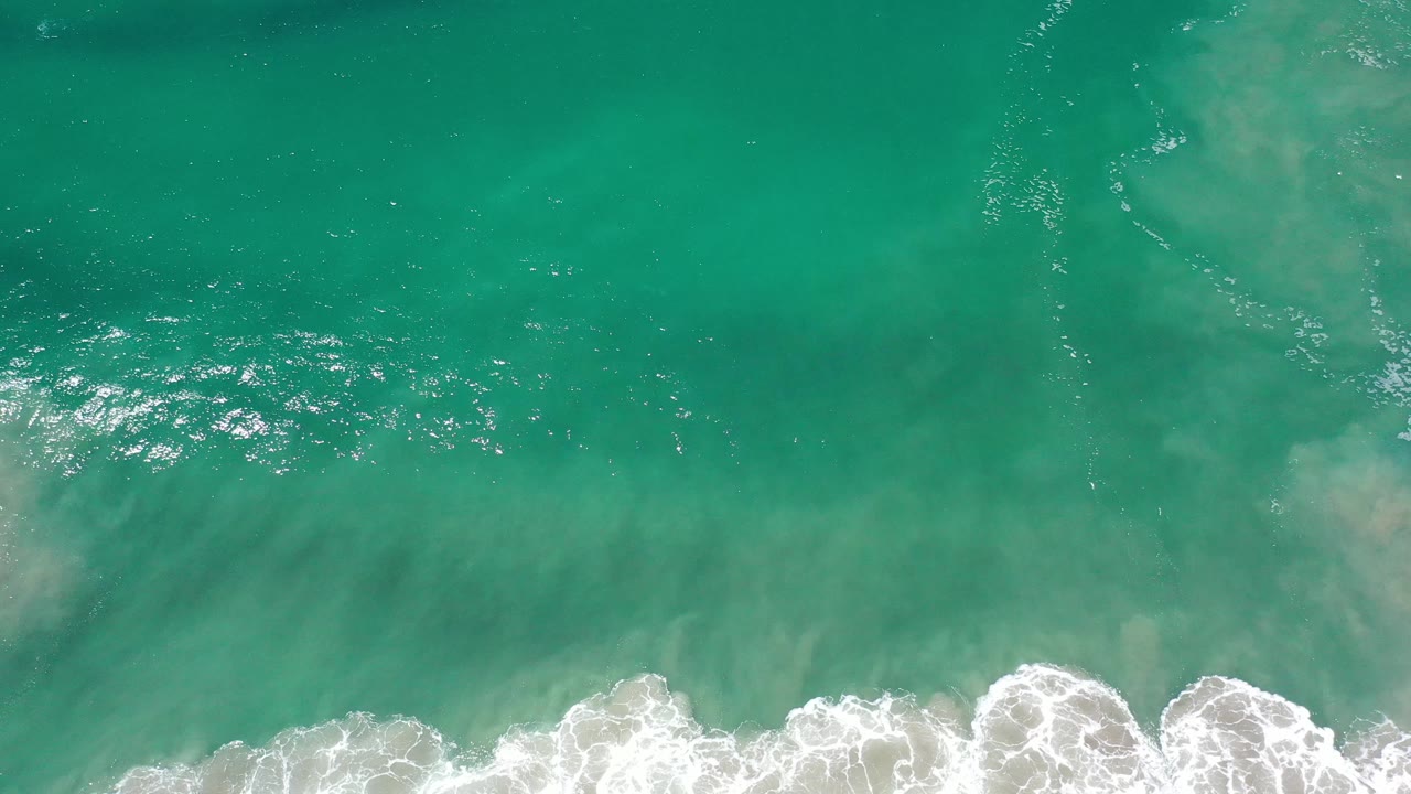 Aerial view of turquoise Los Cabos water with a curling band of white foam near the bottom and no visible people.