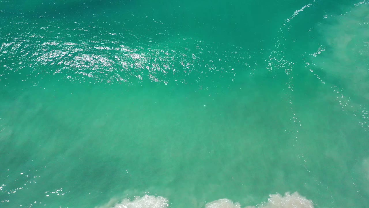 Overhead Los Cabos sea surface showing bright glints near the upper left, soft sand color beneath the water, and no visible people.