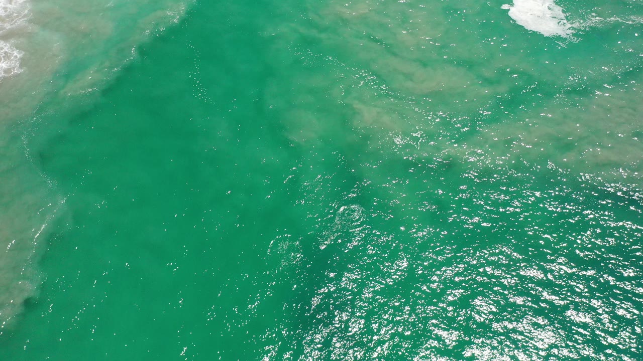 Overhead view of blue-green ocean water in Los Cabos, Mexico, with glittering sunlight, pale wave wash near the top edge, and no visible people.