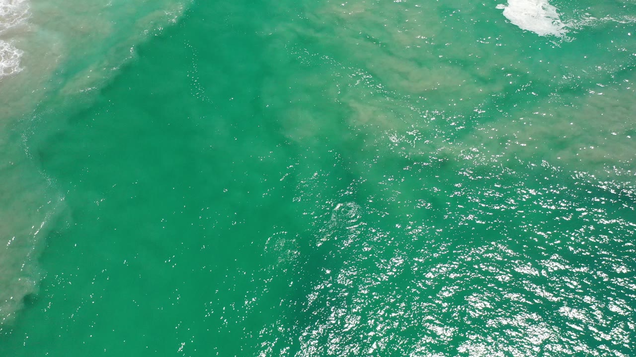 Top-down Los Cabos ocean frame showing turquoise water, faint cloudy sand or foam patterns, bright ripples, and no visible people.