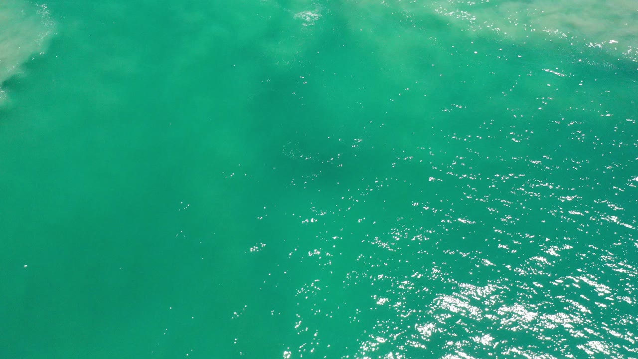 Overhead view of green-blue ocean water in Los Cabos, Mexico, with sunlit ripples and pale cloudy water near the upper edge, no visible people.