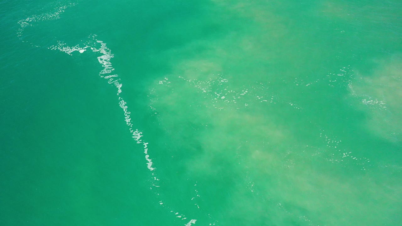 Top-down view of green sea water in Los Cabos, Mexico, with a curved white foam streak along the left side and no visible people.