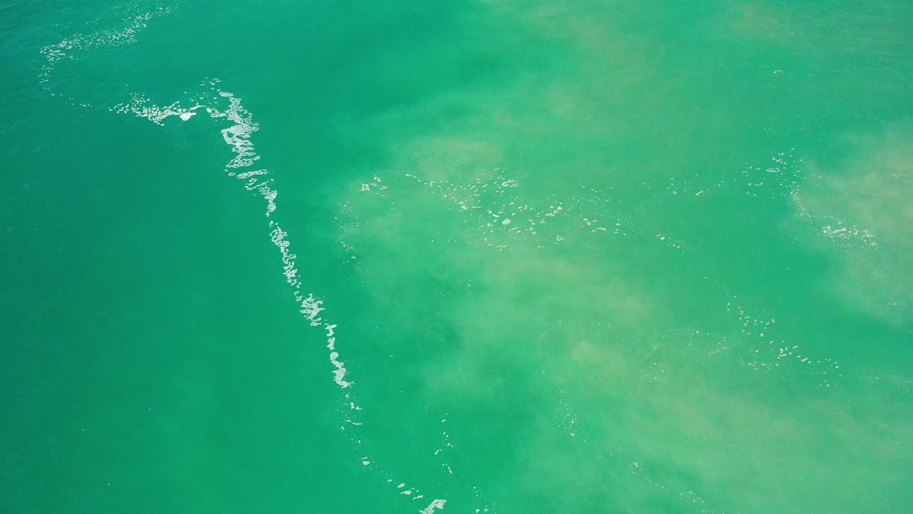 Overhead Los Cabos sea water shows a vertical broken foam line near the left center, pale green underwater patches, and no visible people.