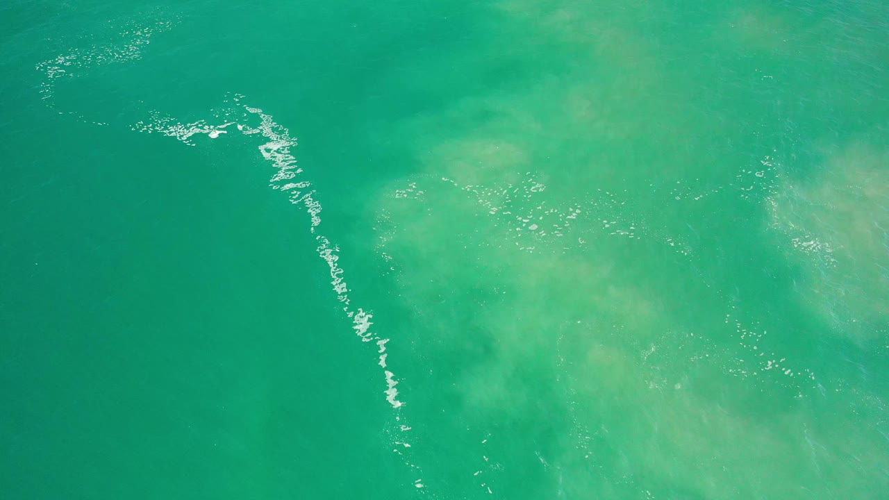 Aerial view over green Los Cabos sea water, where the white foam line sits farther toward the left edge and pale cloudy patches fill the center, with no visible people.