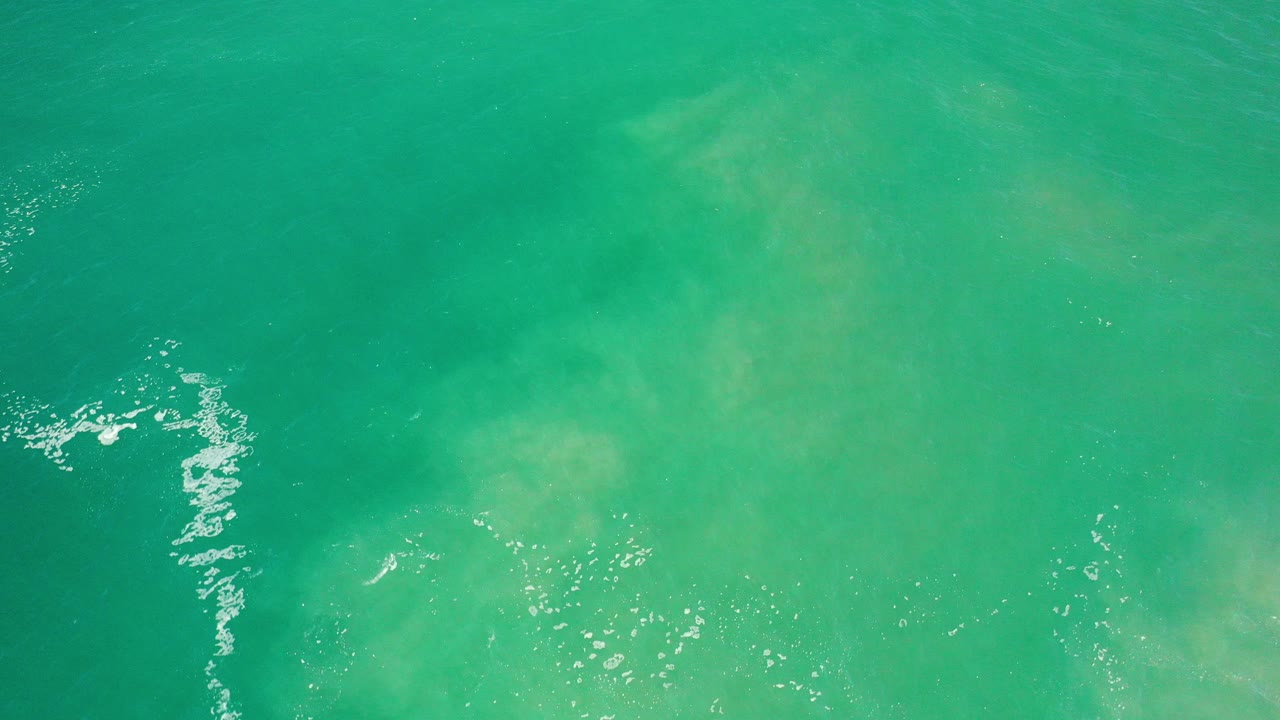 Overhead view of green teal ocean water in Los Cabos, Mexico, with faint subsurface clouding, small white foam at the left edge, and no visible people.