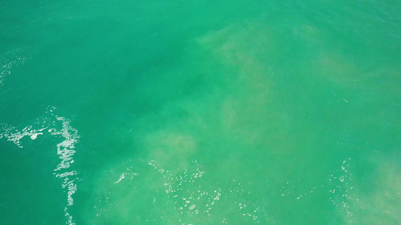 Overhead Los Cabos ocean frame with scattered white wave foam near the lower left, pale green patches under the water, and no visible people.