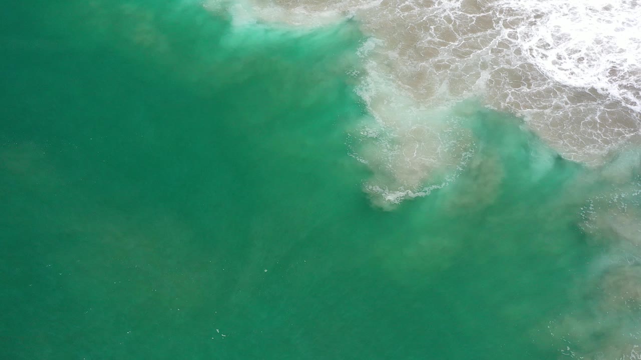 Clear green ocean water in Los Cabos, Mexico, shows a pale sandy or sediment patch beneath the surface below white foam, with no visible people.