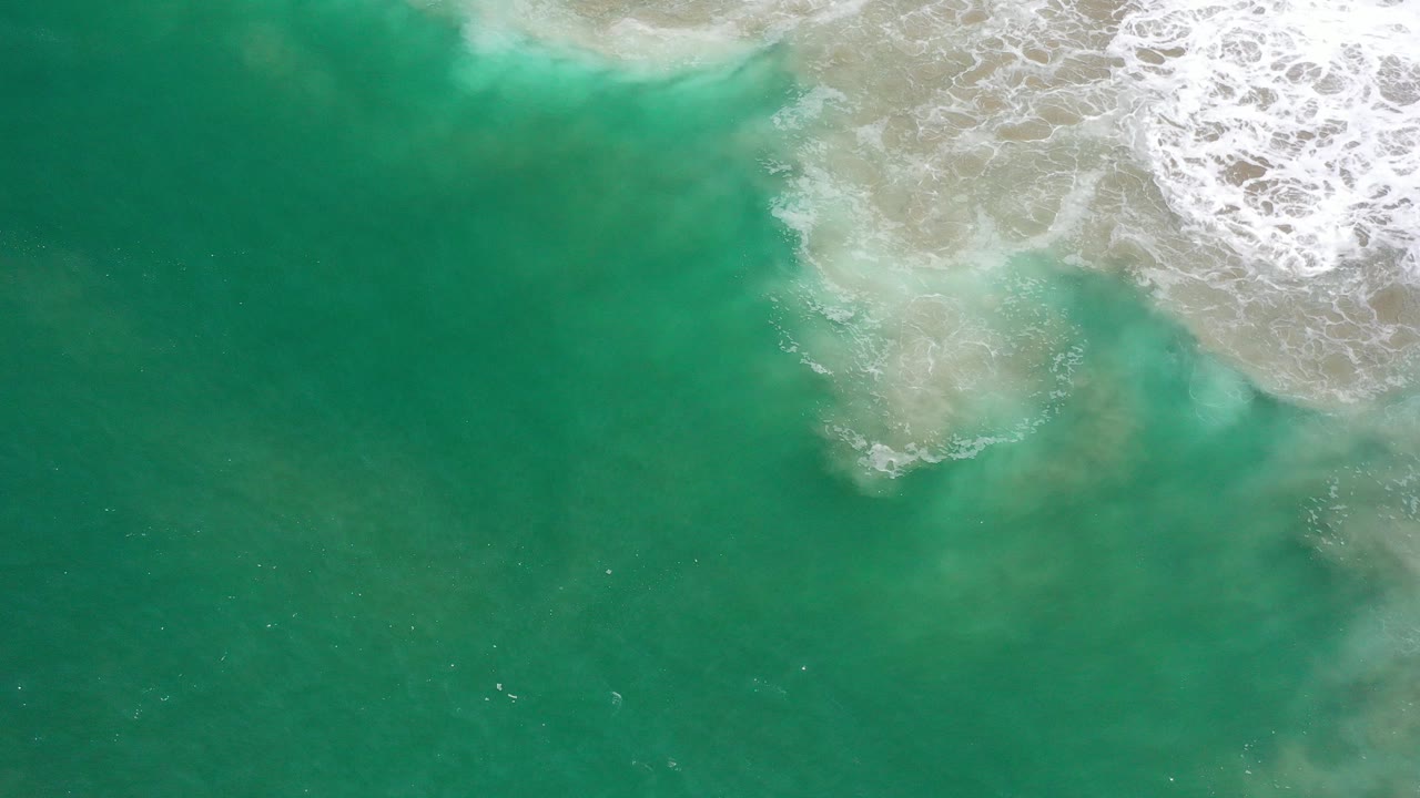 Top-down view of green ocean water in Los Cabos, Mexico, with pale sandy water near the top and white surf foam entering from the upper right, no visible people.