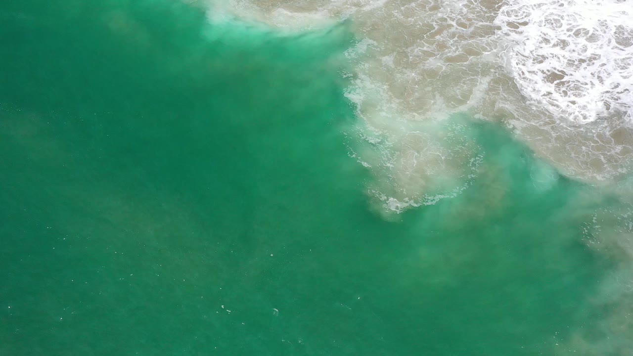 Green sea surface in Los Cabos, Mexico, seen from above as a foamy wave band spreads along the upper edge and cloudy sandy patches sit under the water, no visible people.