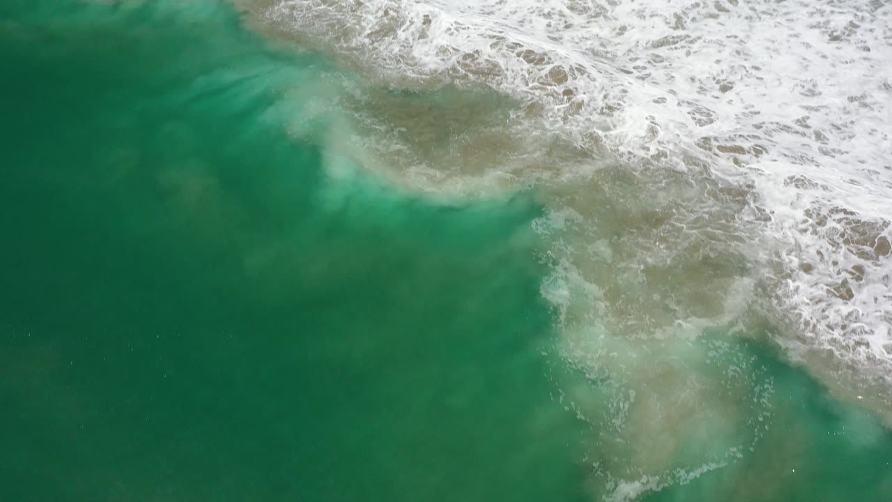 Aerial frame of Los Cabos shoreline water where pale submerged sand shows beneath green water and white foam gathers along the top right, with no visible people.