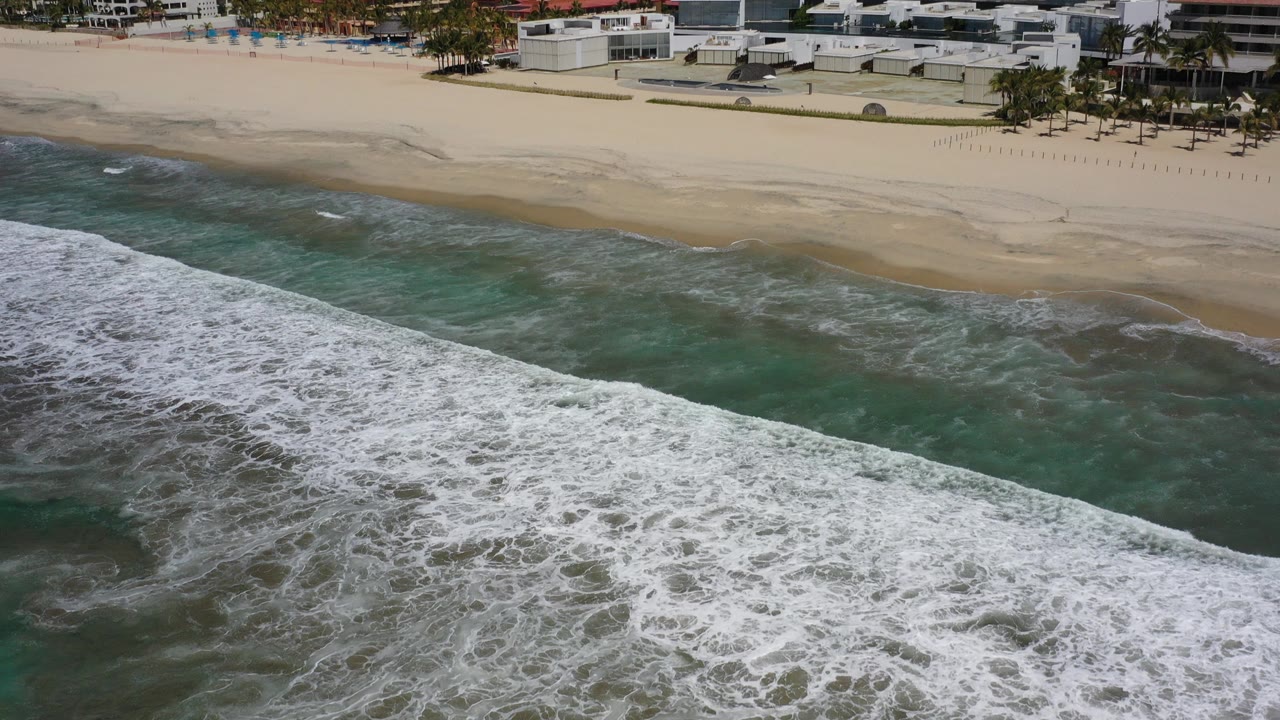 Opening aerial frame of Los Cabos, Mexico, with green water and a wide white foam line below pale sand and beachfront buildings, with no visible people.