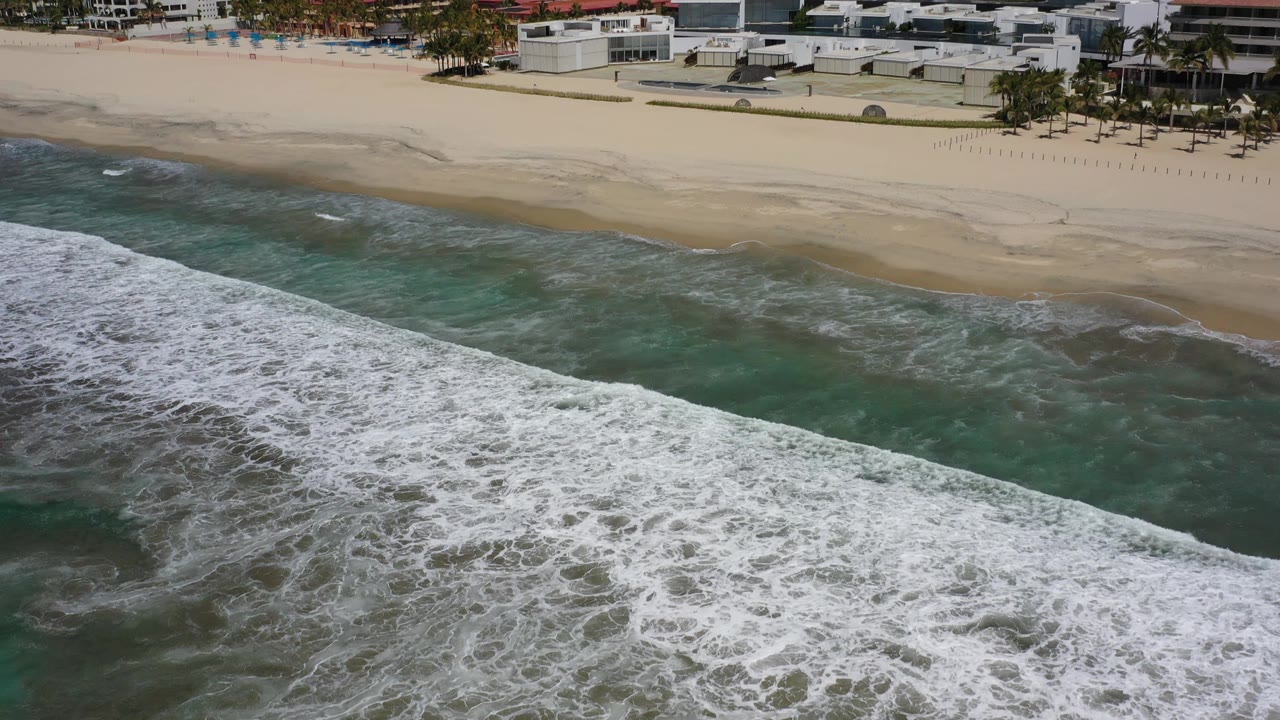 Aerial view in Los Cabos, Mexico, where the white foam line sits lower in frame and shallow green water meets a broad sandy beach, with no visible people.