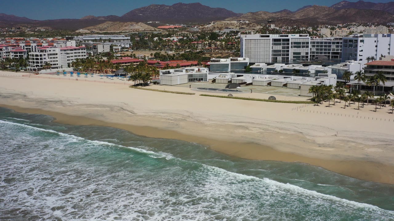 Opening aerial frame over Los Cabos, Mexico, with foamy surf in the foreground, a broad empty beach, white beachfront buildings, palms, and mountains behind the city with no visible people.