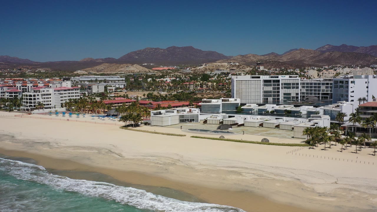Opening frame shows a Los Cabos beachfront from above, with green-blue surf at the lower edge, pale sand, white hotel buildings, distant dry hills, clear sky, and no visible people.