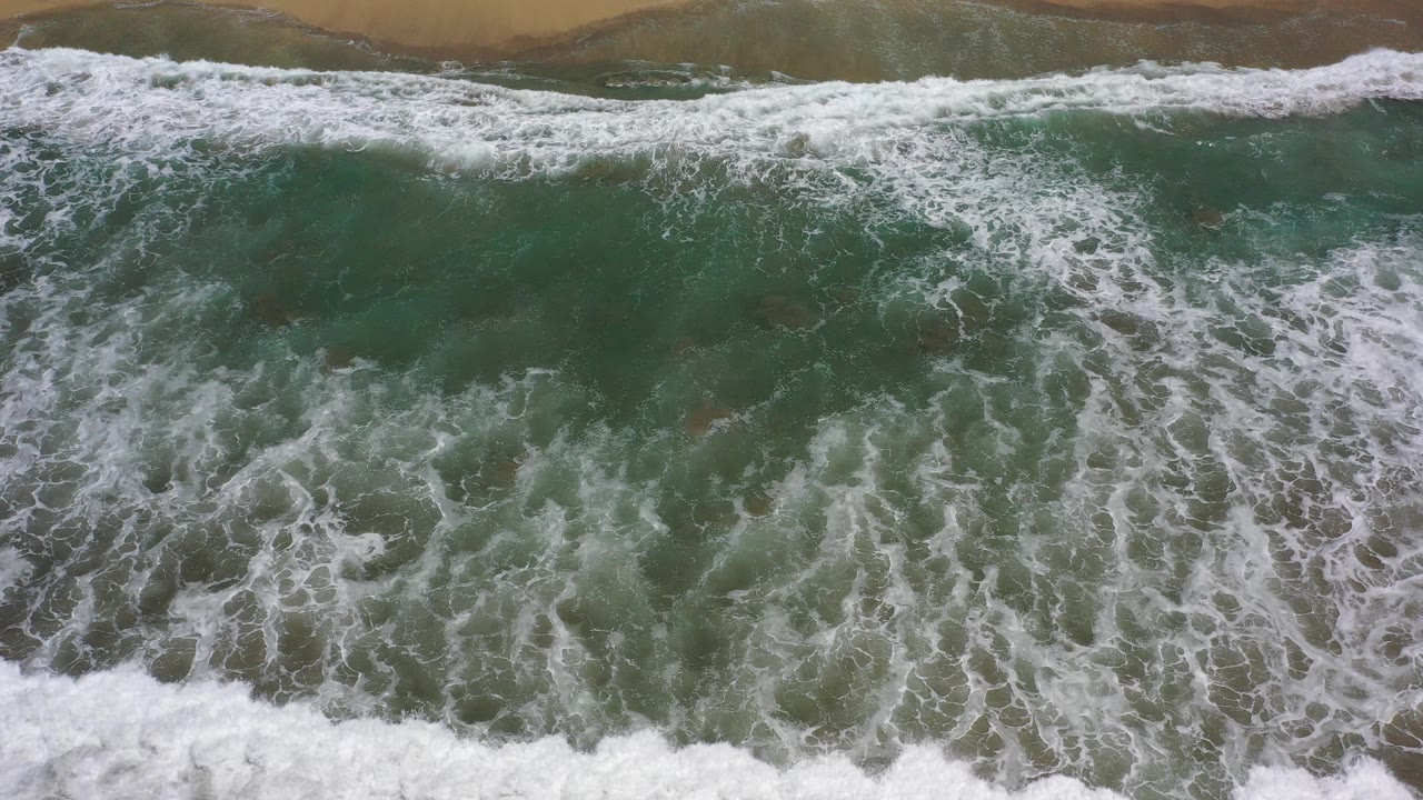 Overhead view in Los Cabos, Mexico, with white surf spreading across green water toward a narrow band of wet dark sand and no visible people.