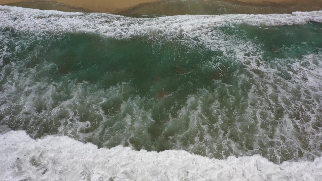 Large foamy wave breaks over green water near dark sand.