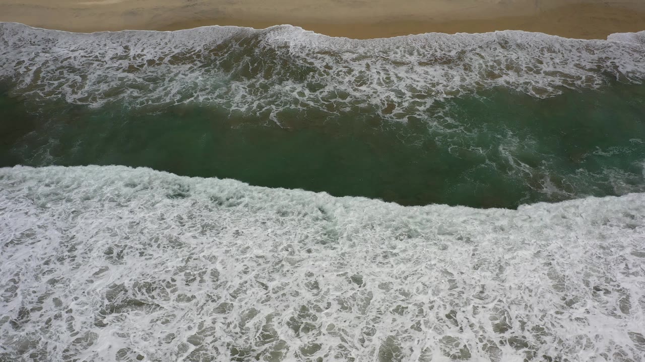Foamy ocean water spreads across a horizontal band of Los Cabos sand as the overhead view stays tight on the shore, with no visible people.