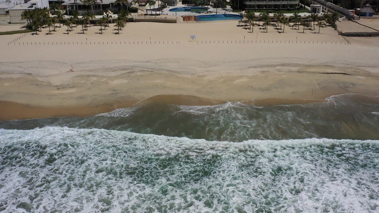 Aerial view in Los Cabos, Mexico, with foamy surf across the lower frame, wet sand in the middle, beachfront hotels and palm trees above, and no visible people.