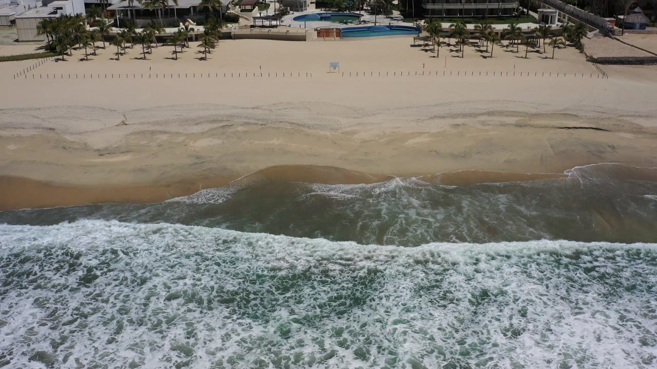 Aerial Los Cabos shoreline view with the white wave band lower in frame, pale sand exposed above it, resort buildings and a blue pool beyond the beach, and no visible people.