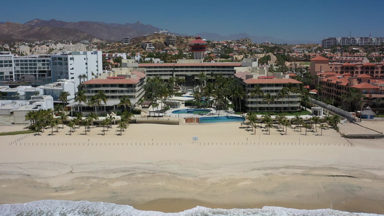 Wide aerial frame over a Los Cabos beachfront, with white surf at the bottom edge, pale sand, resort buildings and palms across the middle, mountains behind, and no visible people.