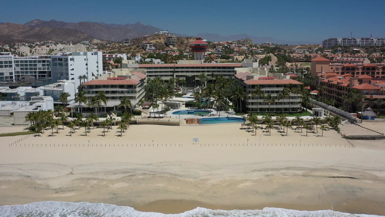 A similar wide Los Cabos aerial view shows the wave line slightly farther up the beach, with pool terraces, palms, low-rise buildings, and no visible people.