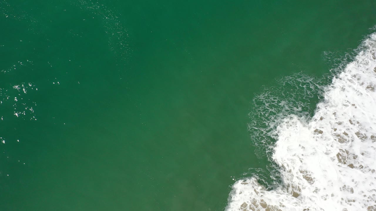 Overhead view of green ocean water in Los Cabos, Mexico, with white foaming surf pushing in from the right side and no visible people.