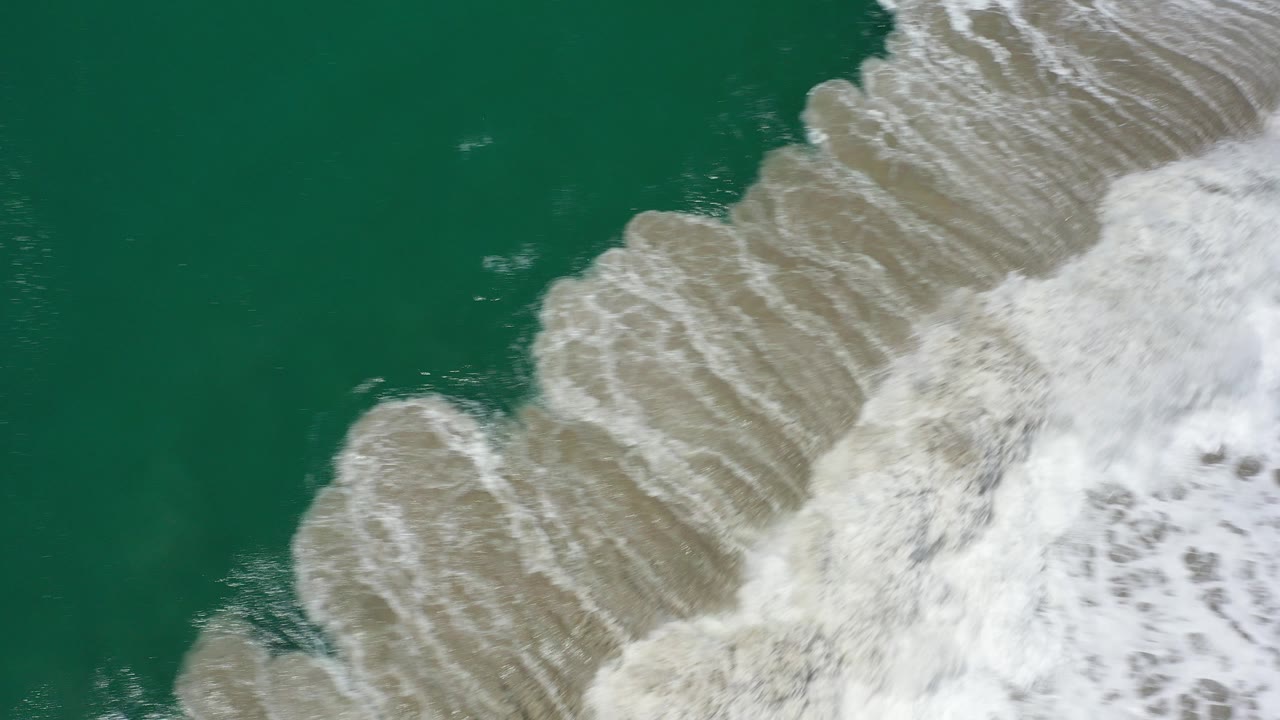 Overhead view in Los Cabos, Mexico, with green ocean water on the left and pale white surf bands breaking diagonally over tan shoreline, no visible people.