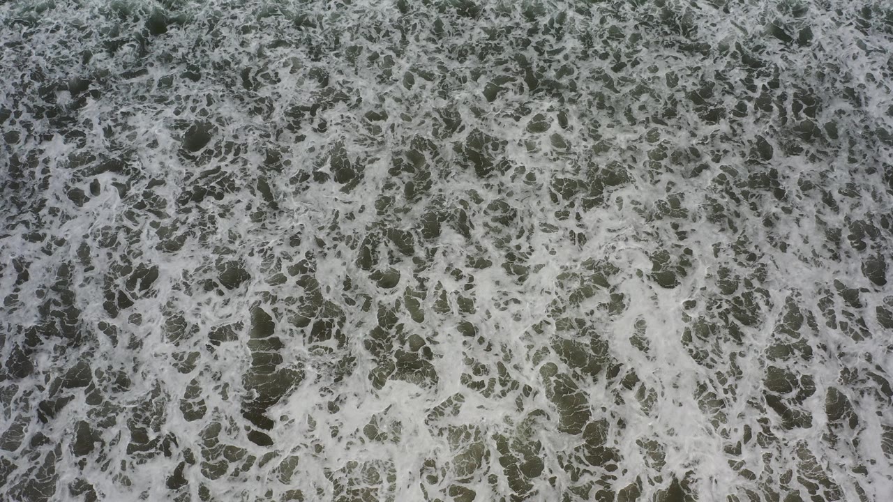 Overhead view of foamy surf over green-gray water in Los Cabos, Mexico, with no shoreline and no visible people.