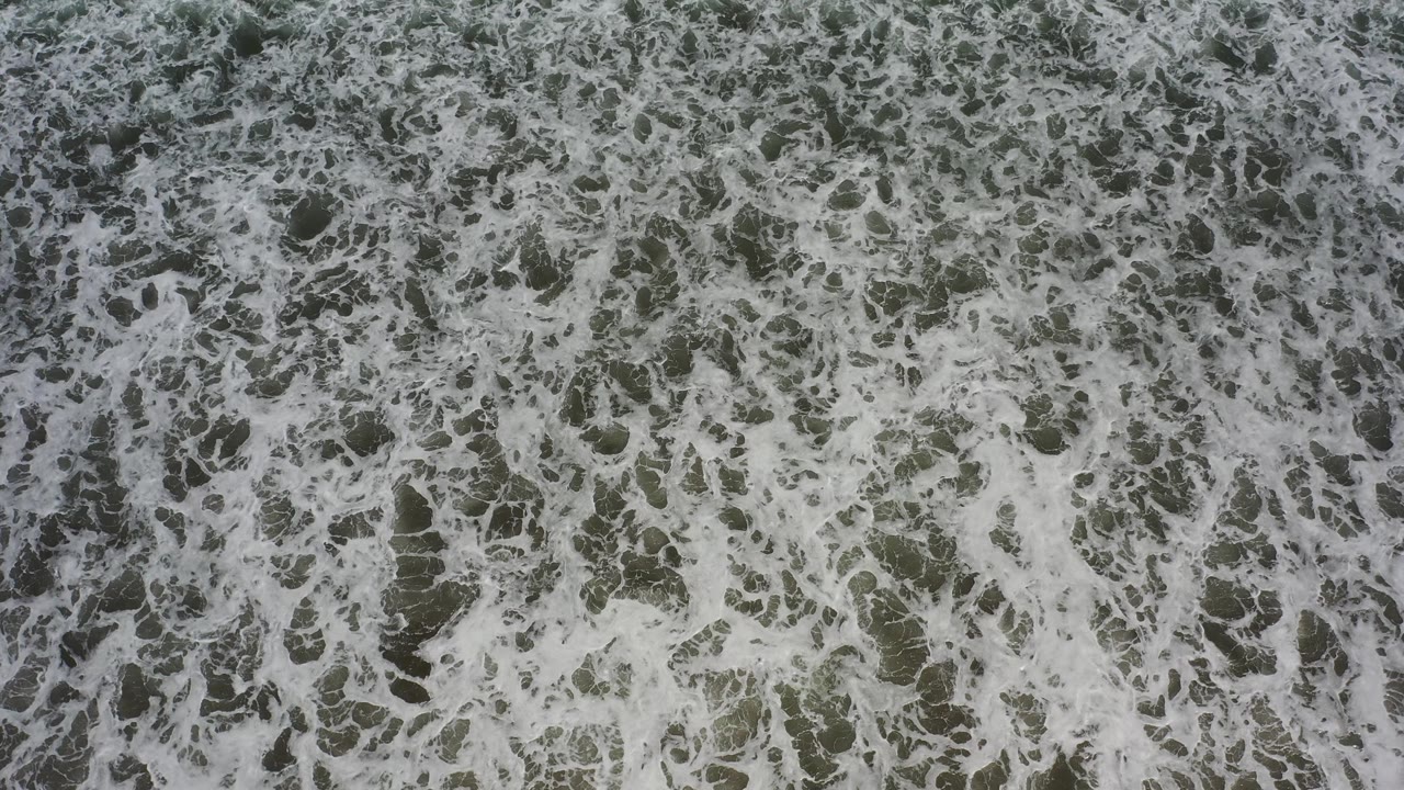 Aerial frame of dense white surf foam spread across green-gray water in Los Cabos, Mexico, with no visible people.
